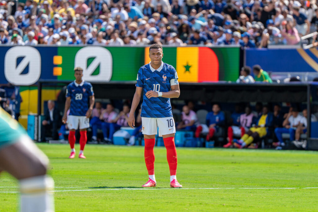 Kylian Mbappé, camisa 10 da França, em pé no gramado durante partida da Copa do Mundo de 2026, com torcida ao fundo.