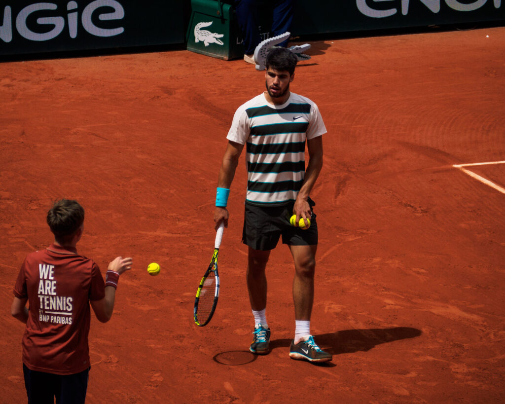 Carlos Alcaraz em quadra de saibro durante partida em Roland Garros 2025, segurando a raquete e recebendo bolas do gandula