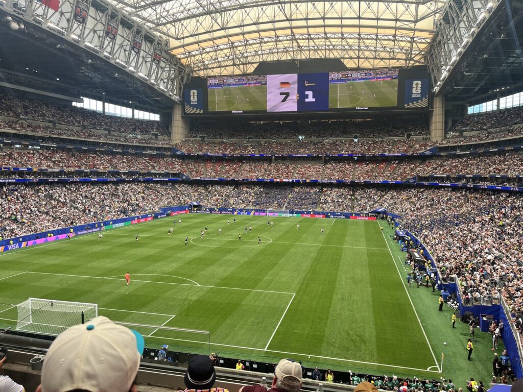 Estádio lotado durante partida da Copa do Mundo de 2026, com jogadores em campo e telão mostrando o placar