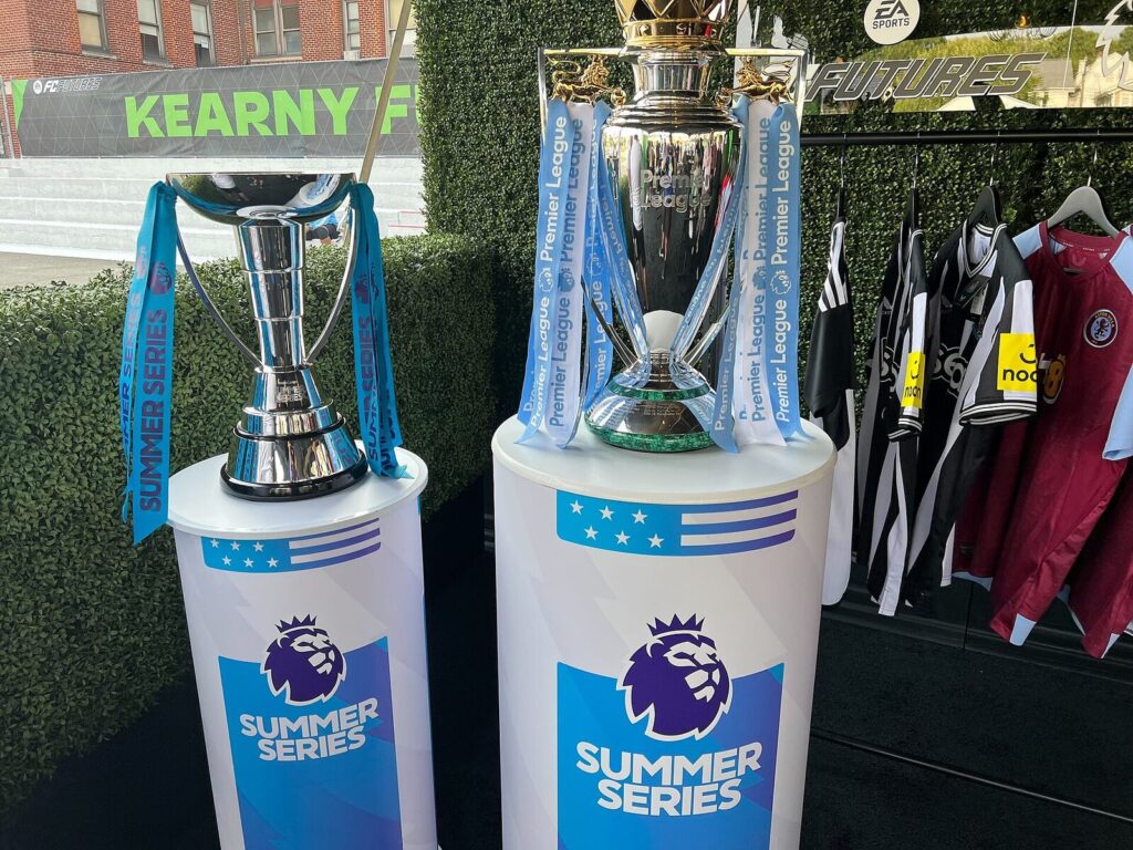 Troféus da Premier League e da Premier League Summer Series expostos lado a lado, com camisas de clubes ao fundo (Foto de arquivo: troféus da Premier League em evento promocional (Summer Series, EUA), sem relação direta com o confronto específico.)