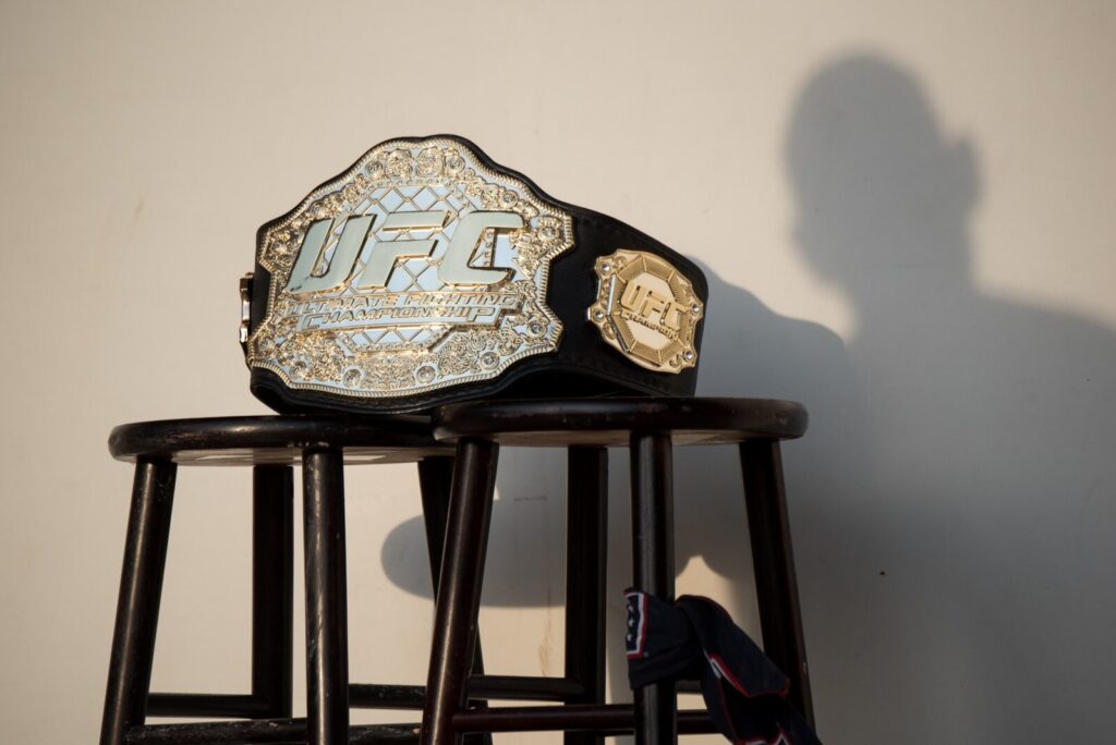 Cinturão de campeão do UFC apoiado em banquetas de madeira (Foto de arquivo: cinturão do UFC exibido em evento de 2018, usado aqui apenas de forma ilustrativa sobre as categorias de peso.)