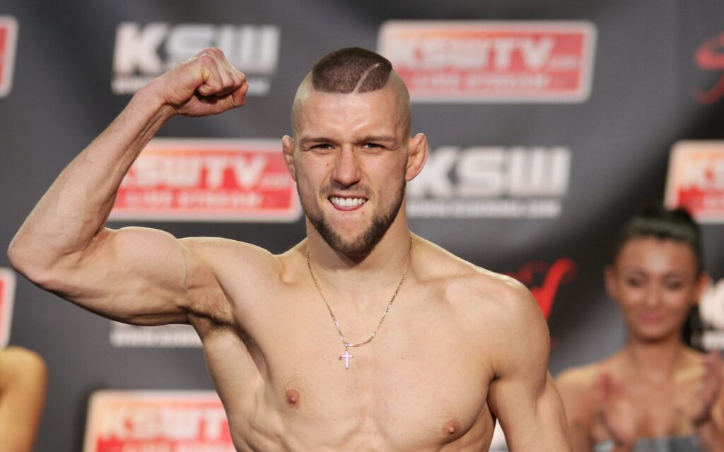 Mateusz Gamrot, lutador polonês, sorrindo e flexionando o braço durante pesagem de evento do KSW (MMA), fundo com logos KSW/KSWTV (Foto de arquivo: Mateusz Gamrot durante pesagem de evento do KSW em 2015, não do UFC Fight Night contra Salkilld.)