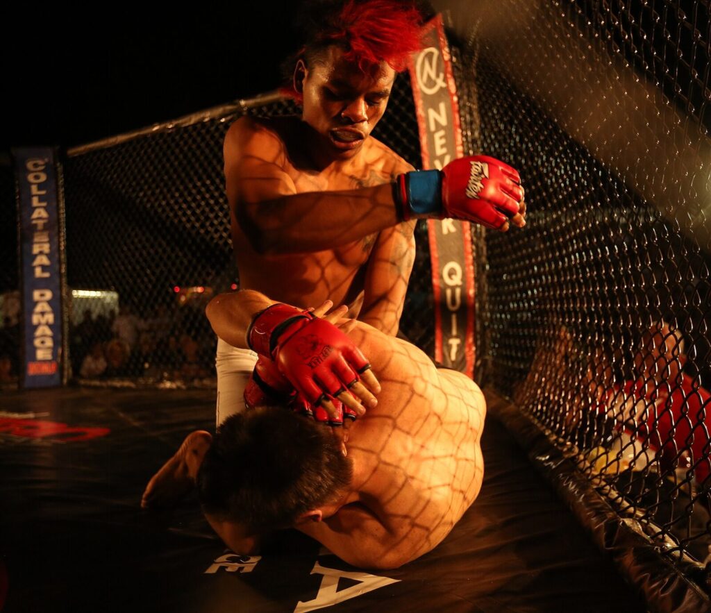 Dois lutadores de MMA durante luta amadora em octógono, um aplicando golpe de cima em posição dominante sobre o oponente no chão (File photo: an amateur MMA cage bout used to illustrate the sport generally, not an actual UFC event or title fight.) (File photo: an amateur MMA cage bout used to illustrate the sport generally, not an actual UFC event or title fight.) (File photo: an amateur MMA cage bout used to illustrate the sport generally, not an actual UFC event or title fight.)