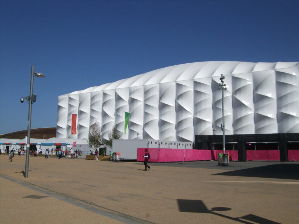 Fachada externa de uma arena de basquete no Parque Olímpico de Londres, com pessoas caminhando em frente sob céu azul (Imagem ilustrativa: arena de basquete no Parque Olímpico de Londres (2012), sem relação direta com o texto.)