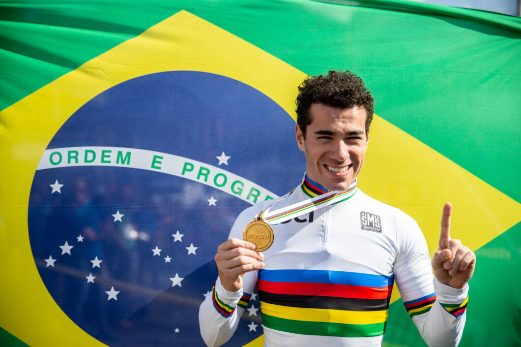 Henrique Avancini sorridente, segurando medalha de ouro, vestindo camisa arco-íris de campeão mundial diante da bandeira do Brasil (Foto de arquivo: Avancini como campeão mundial de MTB Marathon em 2018, não da prova de estrada de hoje em Montreal.)