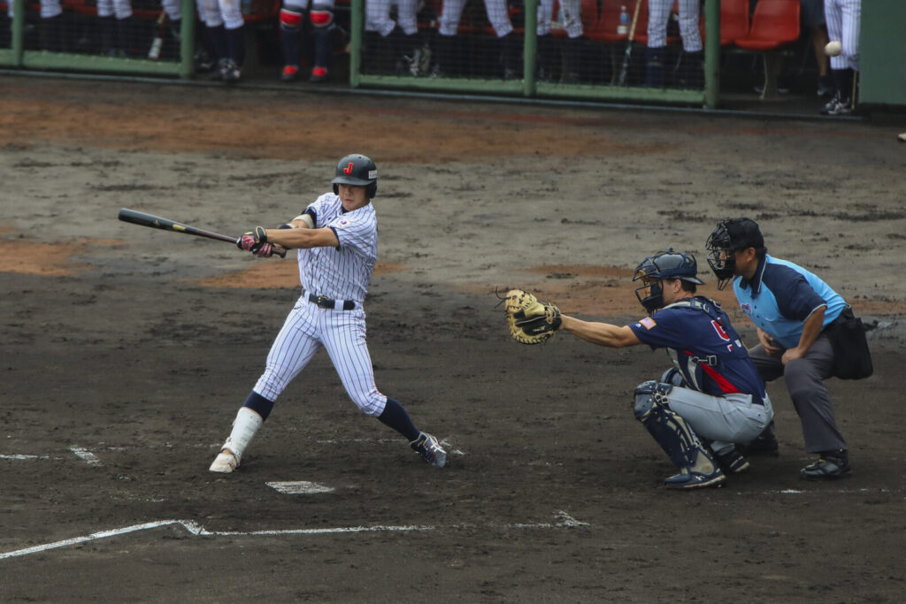 Rebatedor em ação durante partida de beisebol, com apanhador e árbitro na jogada (Foto de arquivo: lance de rebatida no Japan-USA Collegiate Baseball Championship, em Iwakuni (Japão), 2019.)