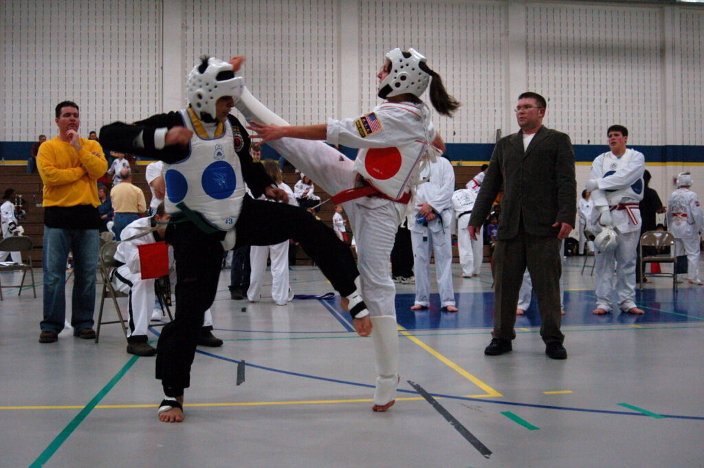 Dois atletas de taekwondo em combate, um com faixa verde e protetor azul, outro com faixa vermelha e protetor vermelho (Foto de arquivo: combate de taekwondo em competição amadora, sem relação com um evento específico.)