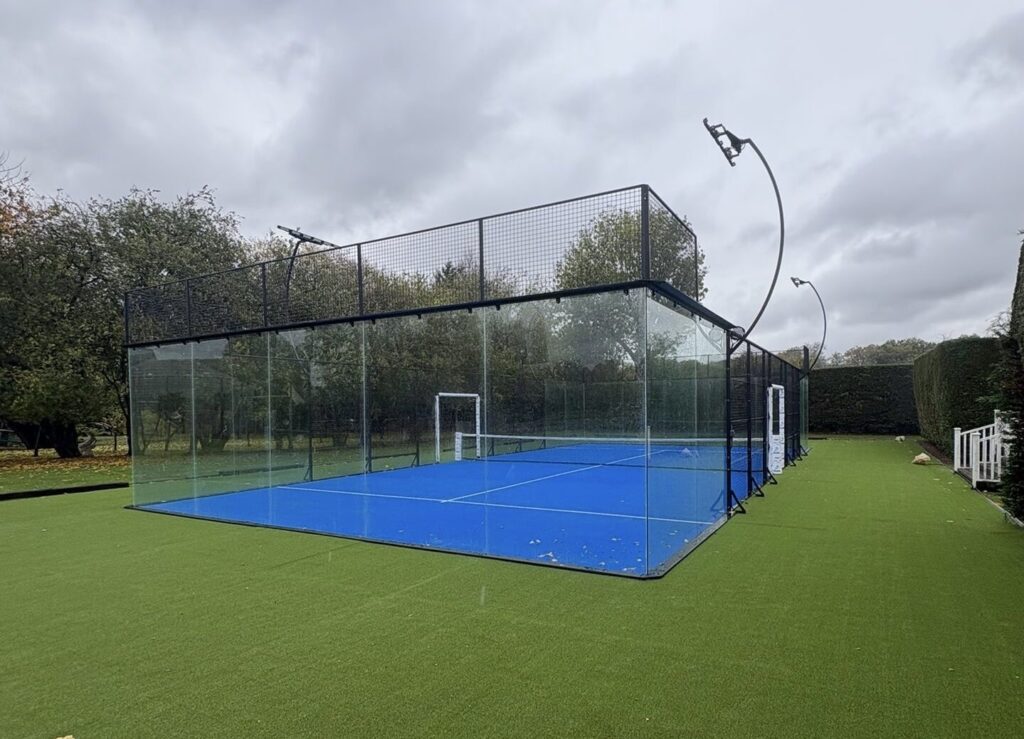 Quadra de padel vazia, com paredes de vidro, grades e piso azul, vista panorâmica sem jogadores (Imagem ilustrativa de uma quadra de padel, sem relação com uma partida específica.)