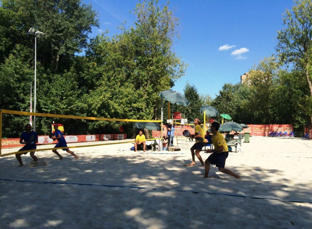 Quatro jogadores disputam partida de beach tennis em quadra de areia com rede, guarda-sóis e público ao fundo (Foto ilustrativa: partida de beach tennis no Campeonato Mundial por Equipes, Rússia, 2014.)