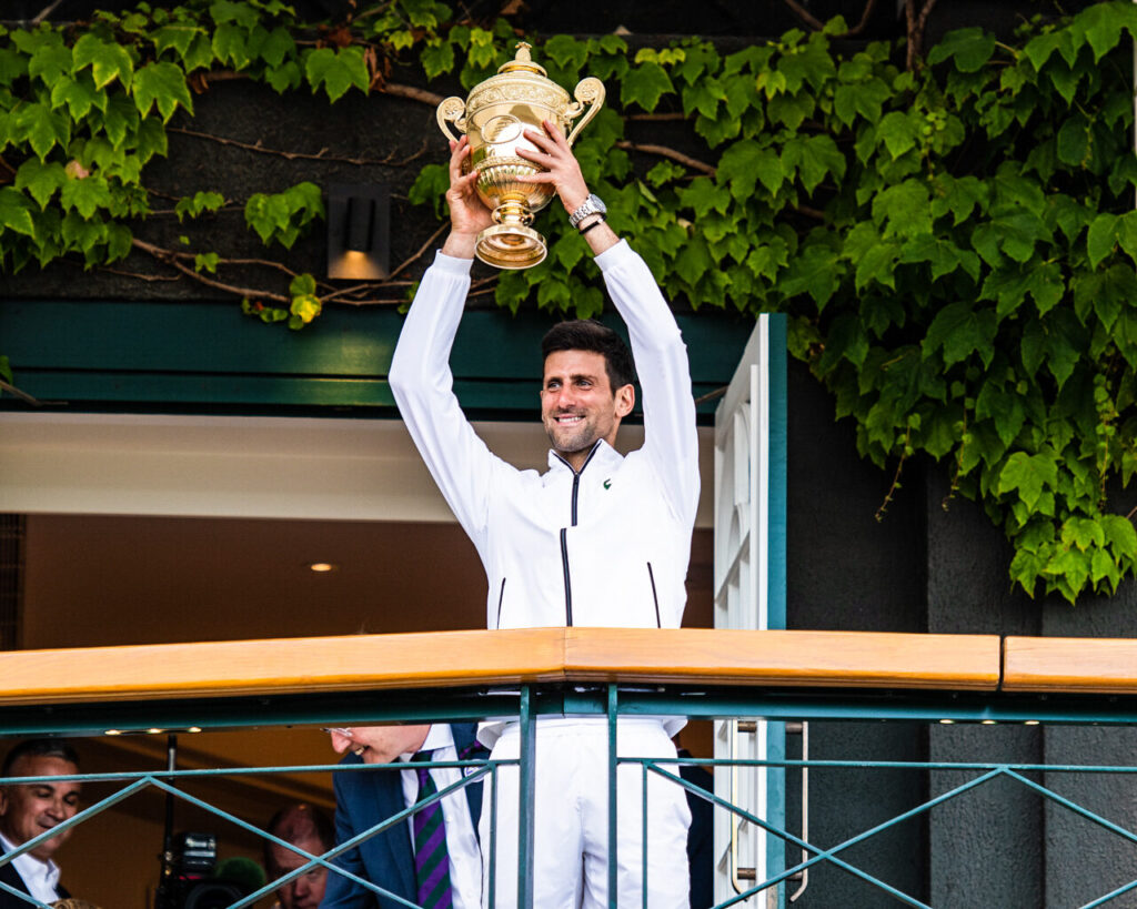 Novak Djokovic ergue o troféu de Wimbledon em 2019, de uniforme branco, sorrindo na varanda do clube (Foto de arquivo: Djokovic levanta o troféu de Wimbledon em 2019, não ligada a uma edição específica do ranking atual de Grand Slams.)