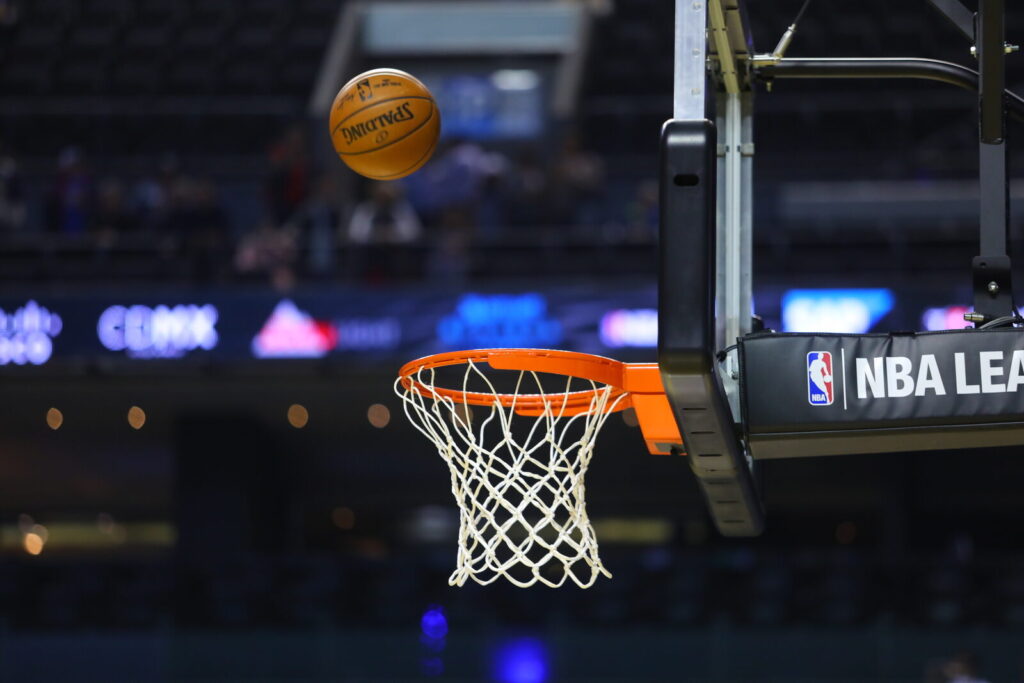 Bola de basquete em direção à cesta com logo da NBA visível na tabela, arquibancada ao fundo (Foto de arquivo: imagem genérica da NBA, sem relação com a estreia específica de LeBron James pelo Sixers.)