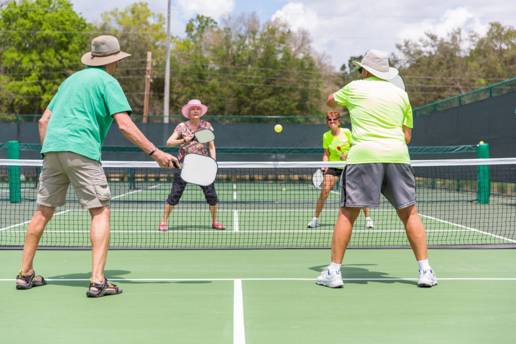 Quatro jogadores em quadra de pickleball durante partida recreativa, com raquetes e bolinha amarela visíveis (Foto de arquivo: partida recreativa de pickleball em The Villages, Flórida.)