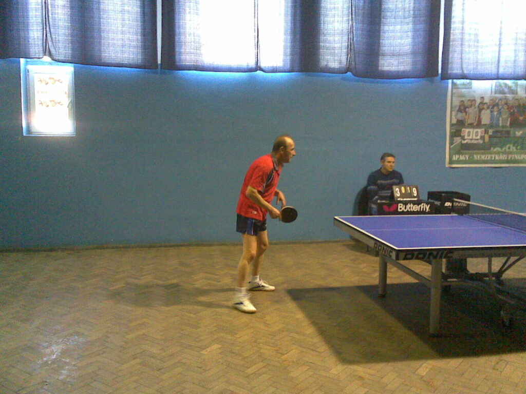 Jogador de camisa vermelha sacando durante partida amadora de tênis de mesa em clube, com adversário e mesa visíveis ao fundo (Foto de arquivo: partida amadora de tênis de mesa, 2009.)